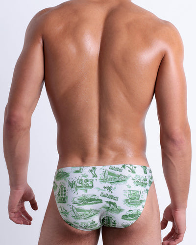 Back view of a model wearing TOILE DE MIAMI (GREEN) men’s beach mini-brief featuring a green and white South Beach, Art Deco building, flamingo, dolphins art made by the Bang! Miami official brand of men's swimwear.