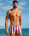 Model wearing The Ken Saint Tropez Swim Shorts featuring bold red, white, and navy vertical stripes, photographed along the shoreline with bright ocean views and coastal scenery.