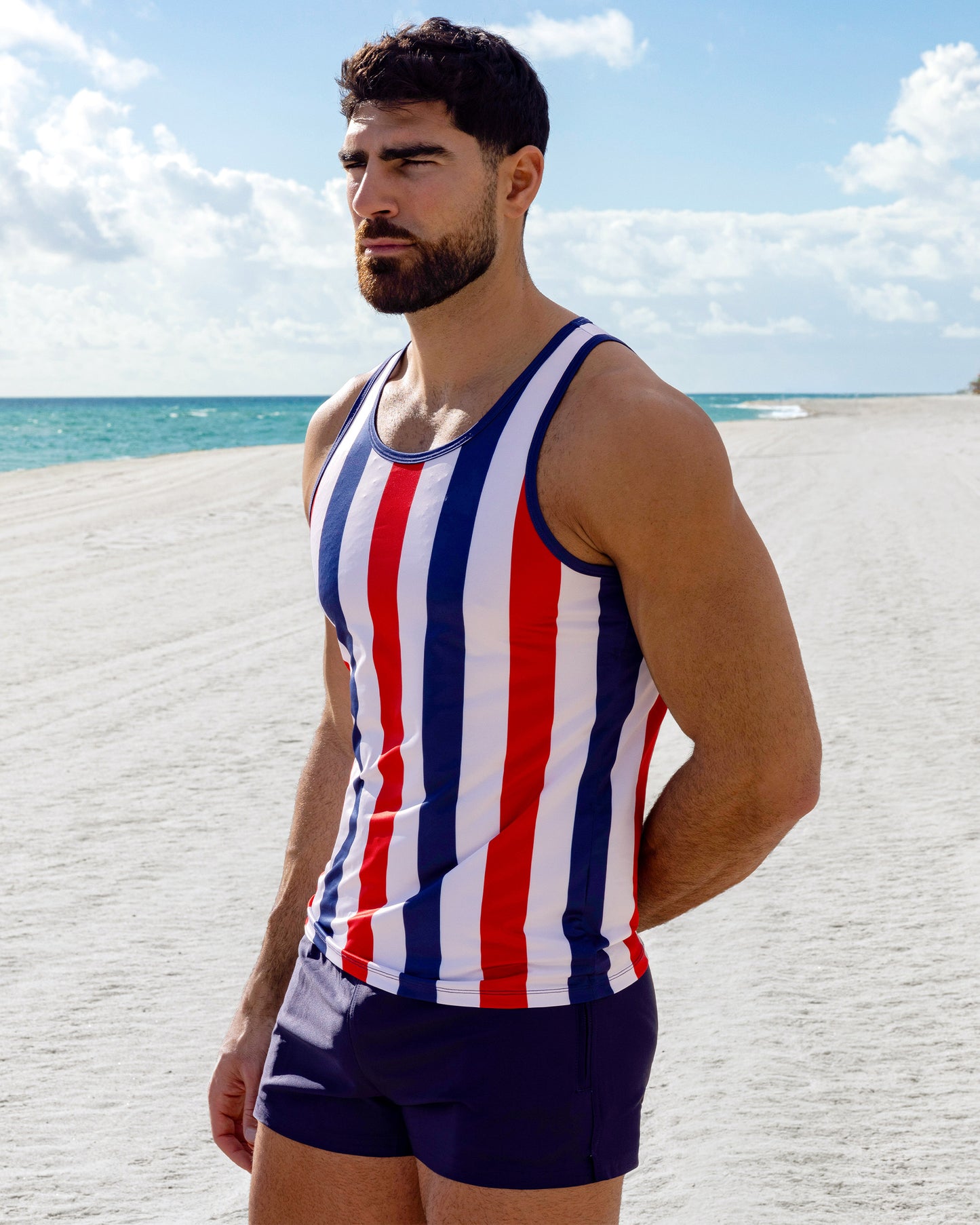 Model wearing The Ken Saint Tropez Tank Top with bold red, white, and navy vertical stripes paired with the Navy Boomer Poolside Shorts, photographed along a bright coastal shoreline.