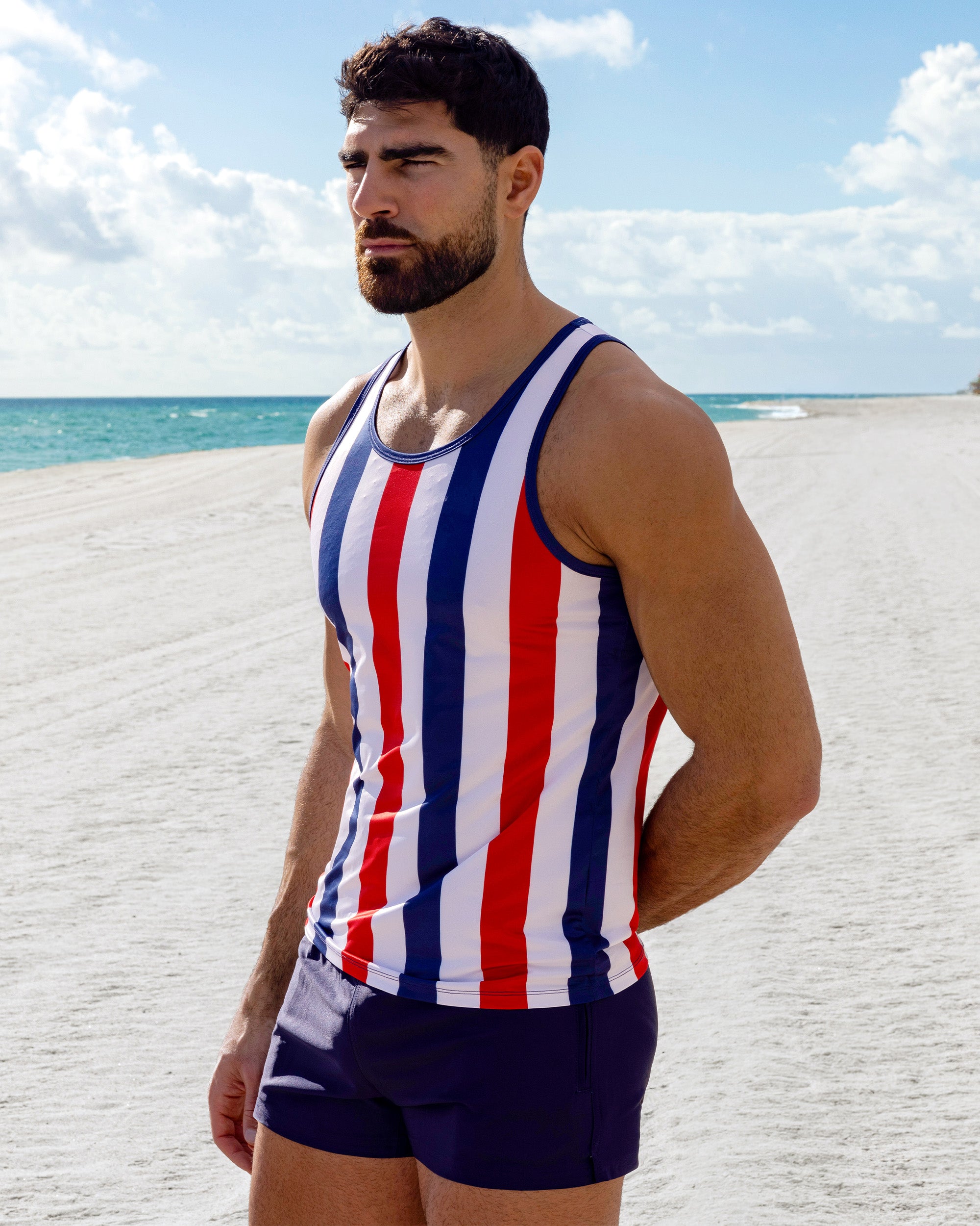 Model wearing The Ken Saint Tropez Tank Top with bold red, white, and navy vertical stripes paired with the Navy Boomer Poolside Shorts, photographed along a bright coastal shoreline.
