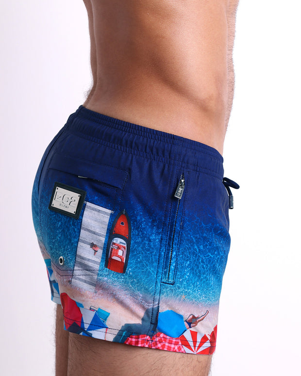 Side view of the UNDER MY UMBRELLA men’s summer Poolside Shorts, with dual zippered pockets. Featuring a photorealistic beach scene print designed by DC2 a brand based in Miami.
