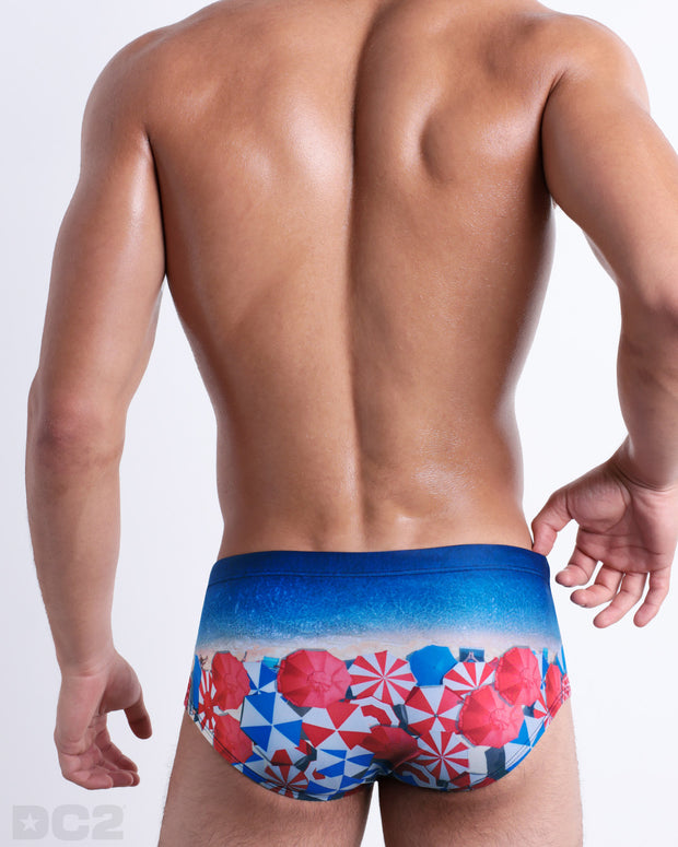 Back view of male model wearing the UNDER MY UMBRELLA beach sexy Brazilian Sunga for men, featuring a photorealistic beach scene print designed by DC2 a brand based in Miami.