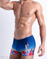 Side view of muscular male model wearing UNDER MY UMBRELLA Summer swimming Trunks. Featuring a photorealistic beach scene print designed by DC2 a brand based in Miami.