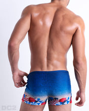 Back view of male model wearing the UNDER MY UMBRELLA beach sexy swimming bottoms. Featuring a photorealistic beach scene print designed by DC2 a brand based in Miami.
