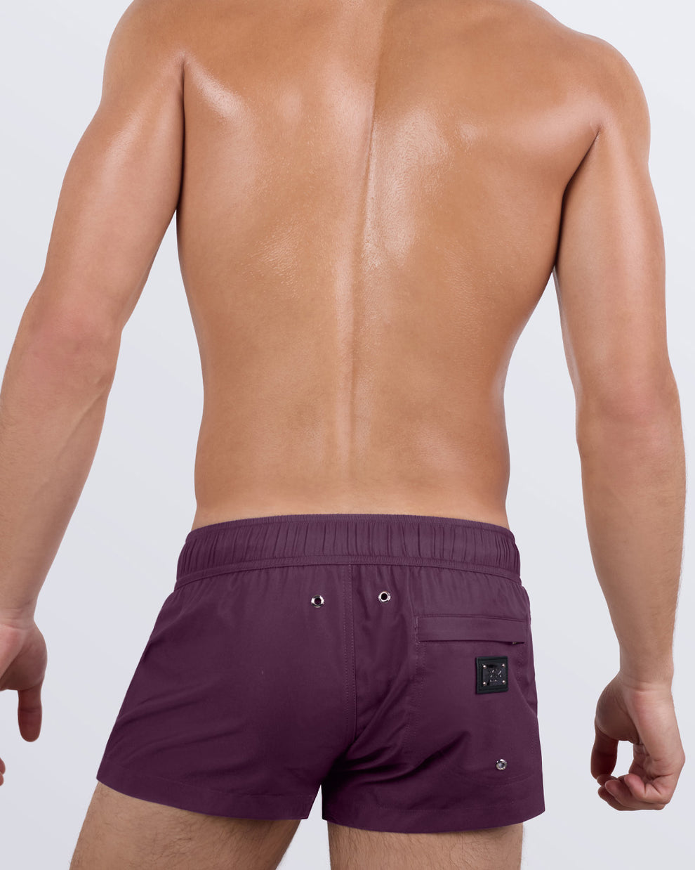 Back view of a male model wearing the men's VERY BERRY Mini Shorts by DC2 Miami. Featuring a deep purple shade, a tailored ultra-short fit, and a functional back pocket with a DC2 signature metal logo plaque, these statement swim shorts offer both comfort and confidence.