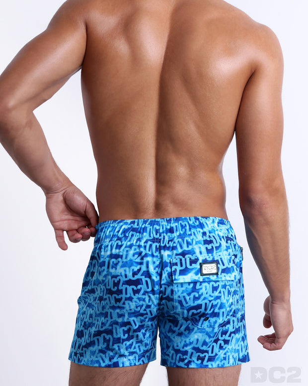 Back view of a male model wearing men’s WET Flex Shorts swimsuits featuring a monogram print of the DC2 logo print designed by DC2 a brand based in Miami.