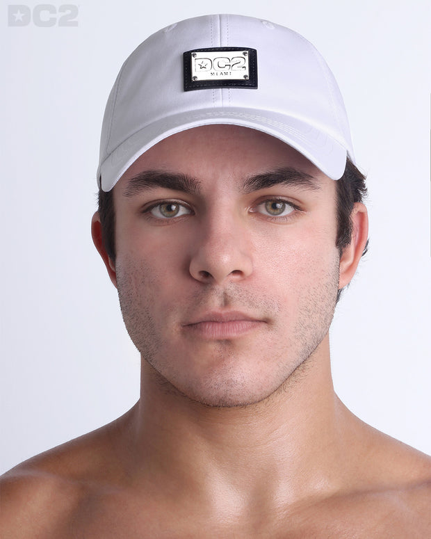 A man wearing the WHITE - Chillax Cap, a stylish white color baseball cap made from breathable fabric. The cap features a metallic silver logo plaque on the front and a curved brim for sun protection.