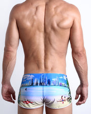 Back view of a male model wearing WISH YOU WERE HERE men’s swim shorts in sky blue with a beach inspired art by the Bang! Clothes brand of men's beachwear.
