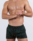 Frontal view of a male model wearing the Written in the Stars Show Shorts for men by BANG! Miami. These premium short-length beach shorts feature a deep black base with an intricate green constellation pattern, an adjustable drawstring waistband with metallic tips, and a tailored fit designed for a bold and confident beachwear look.