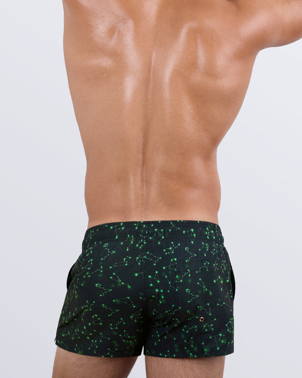 Back view of a male model wearing the Written in the Stars Show Shorts by BANG! Miami. The celestial-inspired beach shorts showcase a functional back pocket with a BANG! logo detail, breathable quick-dry fabric, and a vibrant constellation print wrapping around the entire design.