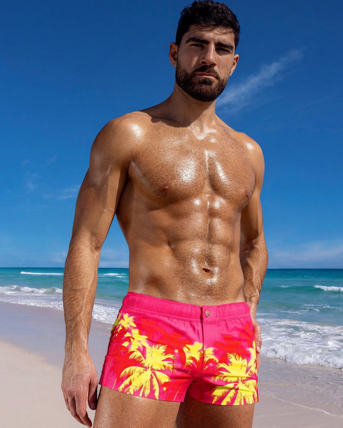 Male model wearing Wuthering Heights (Fucsia) Mini Shorts in a vivid palm print on the beach.
