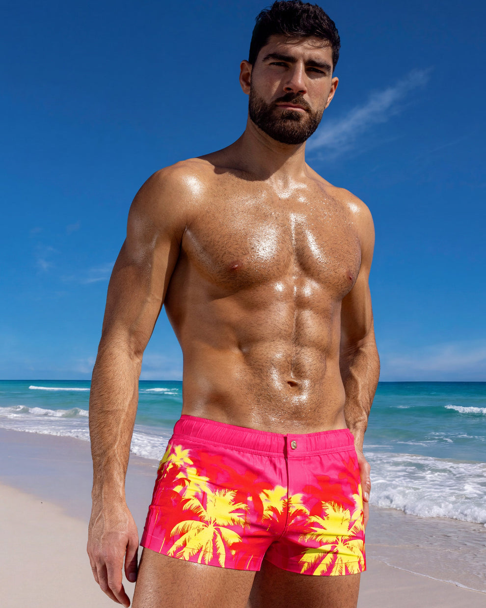 Male model wearing Wuthering Heights (Fucsia) Mini Shorts in a vivid palm print on the beach.
