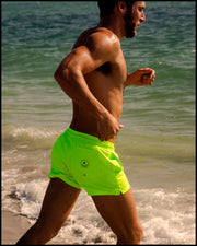 Model wearing men’s swim trunks in neon highlighter yellow color by the Bang! Clothes brand of men's beachwear running at the beach.