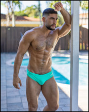Male model wearing the bright AQUA GLOW Swim Brief by a pool outdoors. This swimsuit is quick dry and high-performance with moisture wicking properties.