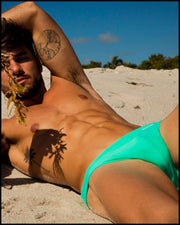 View of a masculine model laying down wearing men’s swim bikini in aquamarine with official logo of BANG! Brand in white.