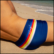 Back view of male model wearing a swim mini-brief in blue color with color stripes in white, yellow, bold red, and blue.