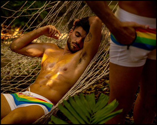 View of male model laying on a hammock wearing the FOUR EVER STRIPES VOL 2 swim trunks for men in solid white with stripes in red, yellow, blue and green by Bang! Clothing of Miami.