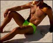 View of a sexy male model laying by the beach wearing men’s swimsuit in bright yellow color by the Bang! Menswear brand from Miami.
