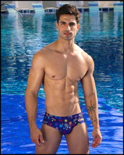 Frontal view of model wearing the HEY MISTER TJ (CLUB MIX) Men’s bikini-style bottoms in dark tones with colorful headphones and disc shapes and a dj tiger print by Bang! men's beachwear.