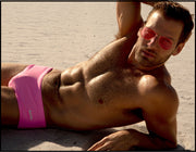 View of a sexy male model laying down by the beach wearing LA BEACH EN ROSE swim sungas in light pink by the Bang! brand of men's beachwear from Miami.