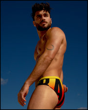 Lateral view of muscular male model wearing BIG BANG GOLDEN HOUR Summer swimsuit for the beach with a bright red, orange and yellow gradient background.