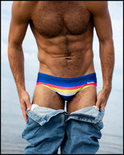 Front view of a sexy male model on the beach wearing a BANG Miami swim mini brief in the classic stripe a pose print