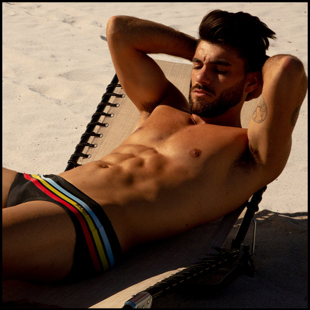 View of model laying out by the beach wearing the BIONIC STRIPES men’s flex boardshorts by the Bang! brand of men&