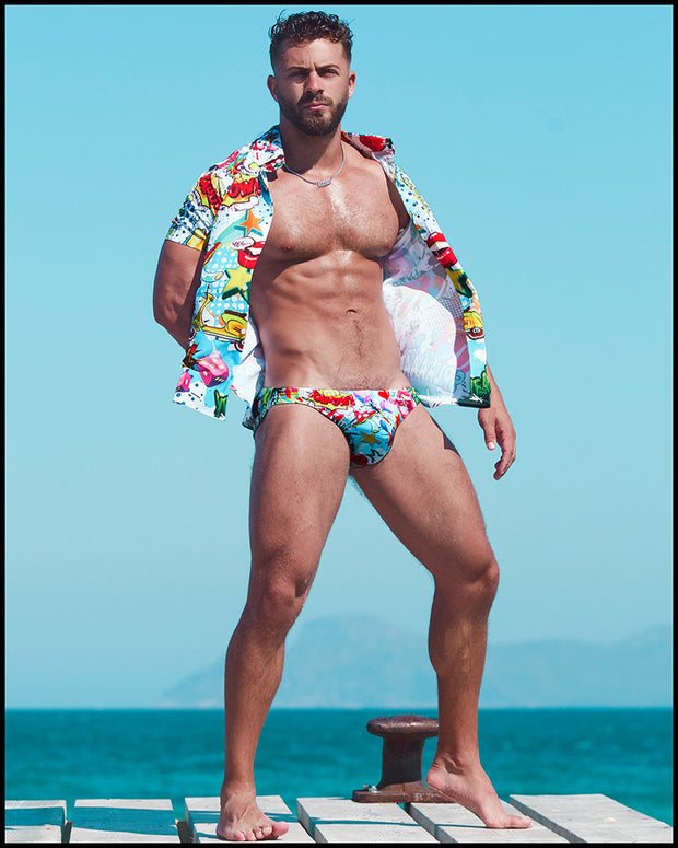 The male model by the ocean wearing the Yeah Yeah swim brief and matching stretch shirt by Bang! Clothes in a cool graffiti print featuring dice and retro cars.