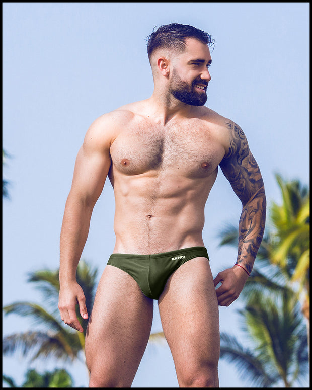 Sexy male model at the beach wearing men’s swimsuit in dark green color by the Bang! Menswear brand from Miami.
