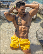 Male model at the beach kneeling down wearing the RADIANT YELLOW Show Shorts by BANG! Clothes the official brand of mens swimwear based in Miami.