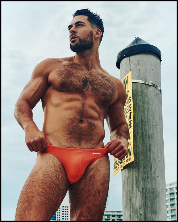 Masculine model outdoors wearing his men’s swimsuit in orange apricot color featuring with official logo of BANG! Brand.
