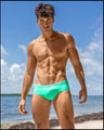 Sultry male model at the beach wearing the AQUA GLOW Swim Brief made with Italian-made Vita By Carvico Econyl Nylon a bright aqua green color. High-performance fabric with moisture wicking properties perfect for a beach day.