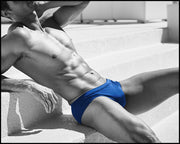 Masculine model sitting down on the stairs wearing men’s swim European bikini in sapphire lining with official logo of BANG! Brand in white.