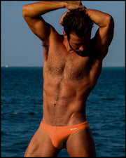 Sexy male model wearing the BRONZE FACTOR a solid orange color Swim Mini Brief outdoors by BANG! Clothes at the beach. 