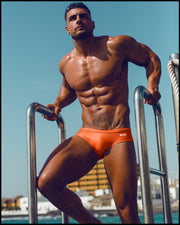 Male model at the beach wearing the BRONZE FACTOR Swim Brief men's swim European bikini in a orange color with the official logo of BANG! Clothes in white.