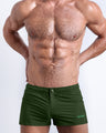 Frontal view of a sexy male model wearing the ALPHA GREEN men’s square leg swim trunks in a solid dark green color by the Bang! Menswear brand from Miami.