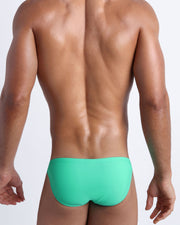 Back view of a male model wearing men’s swim mini-brief made with Italian-made Vita By Carvico Econyl Nylon in aqua color by the Bang! Clothes brand of men's beachwear.