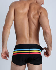 Back side view of male model wearing men's swimwear in black color with color stripes in turquoise blue, yellow, and bold red.
