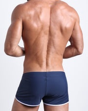 Back view of a male model wearing BLUE JEAN men’s swim shorts in dark navy blue color by the Bang! Clothes brand of men's beachwear.
