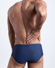 Back view of a male model wearing men’s BLUE JEAN swim sungas in a dark navy blue color by the Bang! Clothes brand of men's beachwear.