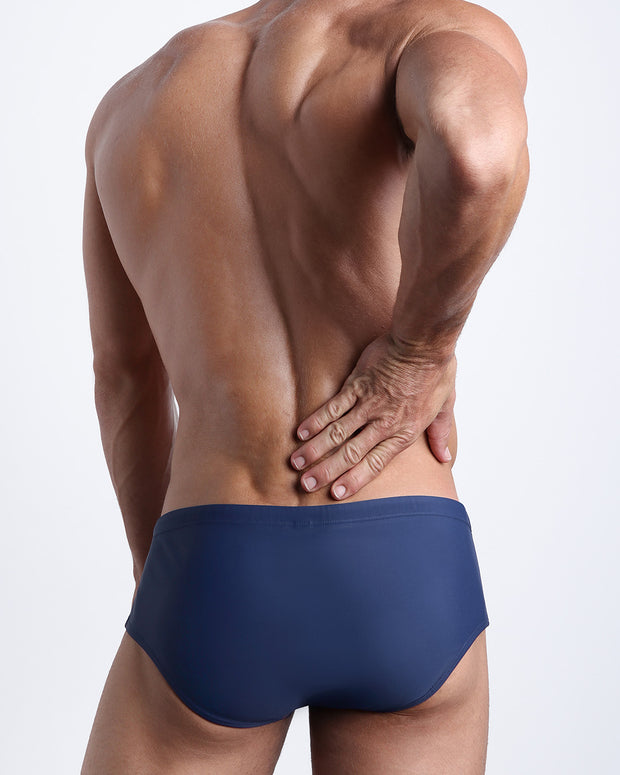 Back view of a male model wearing men’s BLUE JEAN swim sungas in a dark navy blue color by the Bang! Clothes brand of men&