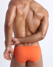Back view of a male model wearing men’s swim briefs in apricot color by the Bang! Clothes brand of men's beachwear.