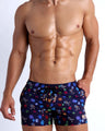Frontal view of model wearing the HEY MISTER TJ (CLUB MIX) Men’s beach shorts in dark tones with colorful headphones and disc shapes and a dj tiger print by Bang! men's beachwear.