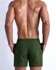 Back view of male model wearing the ALPHA GREEN beach trunks for men by BANG! Miami in army military green. 