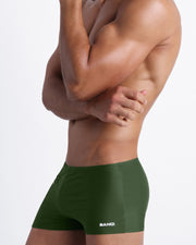 Side view of a masculine model wearing men’s beach swimwear in pine color with official logo of BANG! Brand in white.