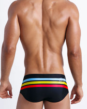 Back side view of sexy male model wearing men's swimwear trunks in black color with color stripes in turquoise blue, yellow, and bold red.