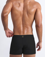 Back view of a masculine model wearing men’s swim summer beach shorts in solid black with official logo of BANG! Brand in gold.