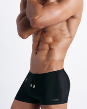 Left side view of a masculine model wearing men’s swimsuit in solid black with official logo of BANG! Brand in gold.