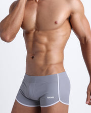Left side view of a masculine model wearing men’s swimwear in silver lining with official logo of BANG! Brand in white.