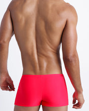 Back view of a sexy male model wearing men’s swim trunks in neon red by the Bang! Clothes brand of men's beachwear.